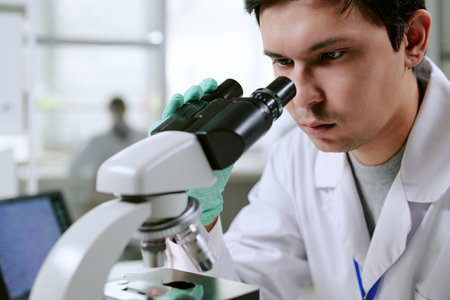 Young adult man examining sample through microscope in laboratory setting, wearing lab coat and gloves, focusing intently on scientific research in medical environmentの写真素材