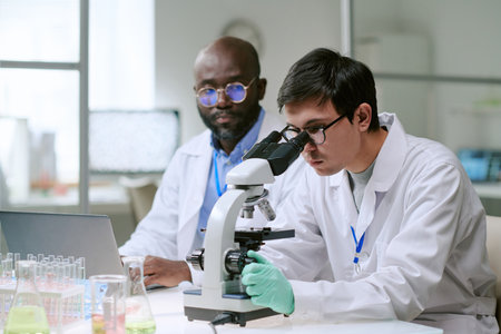 Man examining sample with microscope while another man observing and working on laptop in laboratory setting with scientific glassware visibleの写真素材
