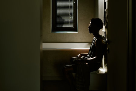 Young adult man sitting on bathtub edge in morning light, gazing thoughtfully toward window, short curly hair visible, relaxed posture, towel draped over lap, minimalist bathroomの写真素材