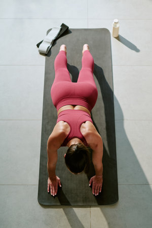 Young adult woman performing plank exercise on yoga mat during morning workout, hair tied back, fitness band and water bottle placed nearby on tiled floorの写真素材