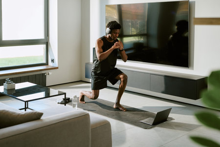Young adult Black man exercising on yoga mat in modern living room, wearing headphones, performing lunge while following workout routine on laptop during morningの写真素材