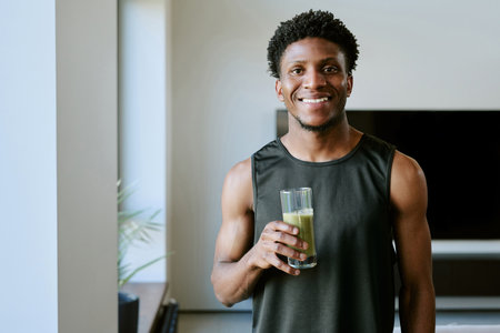 Portrait of young adult Black man smiling and holding glass of green smoothie in modern home setting, standing near window with natural morning light illuminating face and upper bodyの写真素材