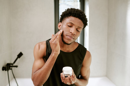Young adult man standing in bathroom applying facial cream to cheek with eyes closed, holding skincare jar in hand, morning beauty routine emphasizing self care and groomingの写真素材