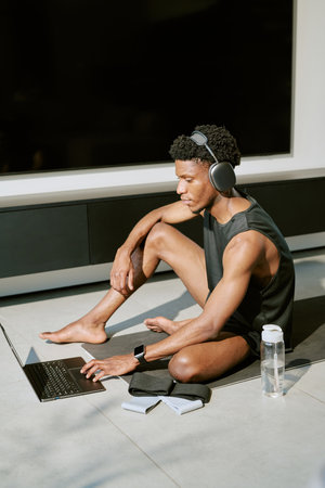 Young adult Black man sitting on floor using laptop and wearing wireless headphones, holding one knee while looking at screen, water bottle and notebook placed nearby, morning light streaming inの写真素材