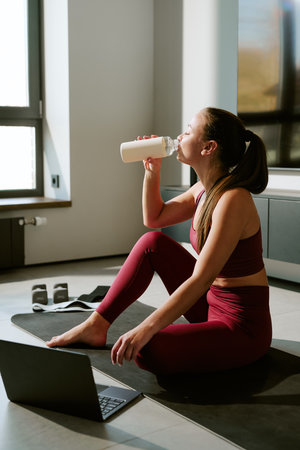 Young adult woman sitting on yoga mat drinking protein shake after morning workout, laptop and...
