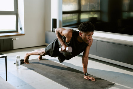 Young adult man exercising on yoga mat performing one arm push up with dumbbell in modern living...