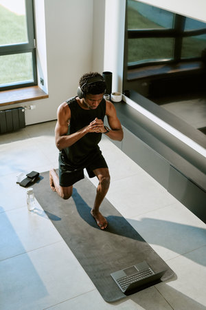 Young adult Black man exercising on yoga mat indoors, wearing headphones and kneeling with hands...