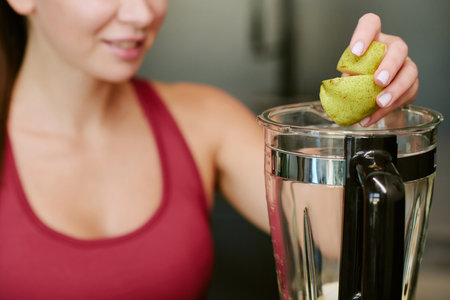 Young adult woman preparing healthy breakfast, smiling while placing fresh pear slices into blender,...