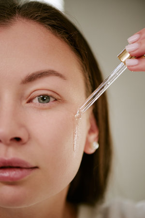 Portrait of young adult Caucasian woman applying facial serum with dropper to cheek, closeup showing skincare routine and healthy complexion, morning beauty conceptの写真素材
