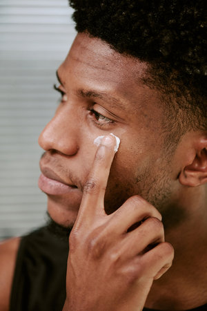 Young adult Black man applying facial cream to cheek with finger, focusing on skincare routine in morning, closeup showing side profile and gentle expressionの写真素材