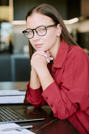 Portrait of young adult Caucasian woman with long straight hair and glasses sitting at desk, resting chin on hands, looking into camera, laptop and documents visible on tableの写真素材
