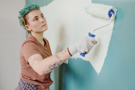 Caucasian young adult woman painting wall with paint roller during home renovation, showing focused...