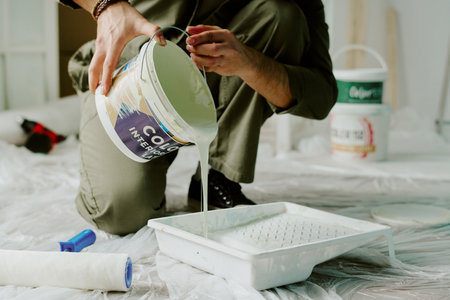 Man pouring paint from large can into paint tray while kneeling on floor during home renovation, plastic sheeting and painting supplies visible aroundの写真素材