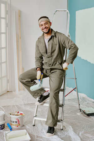 Portrait of young adult man smiling while sitting on ladder holding paint roller, wearing work gloves and coveralls, preparing wall for painting during home renovationの写真素材