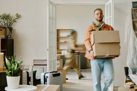 Young adult Caucasian man carrying cardboard boxes with potted plant while young adult woman moving in background, both engaged in home moving and renovation activitiesの写真素材