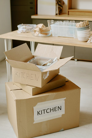 Cardboard boxes labeled kitchen stacked on floor with plastic containers and utensils inside, young couple unpacking and organizing kitchen items during moving and renovation processの写真素材