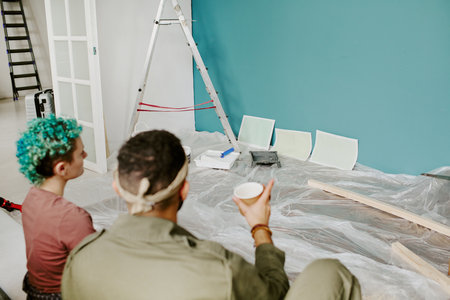 Young adult couple sitting on floor taking break during home renovation, looking at paint color...