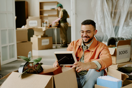 Young adult man sitting among moving boxes smiling while unpacking framed photo, young adult woman...