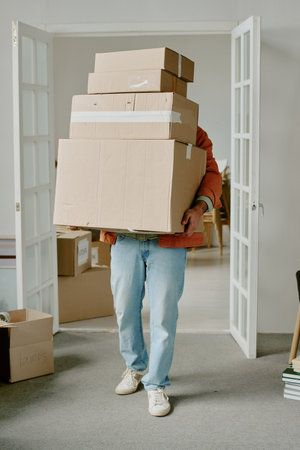 Caucasian young adult man carrying large stack of cardboard boxes while moving into new home, standing in doorway with additional moving boxes visible in backgroundの写真素材