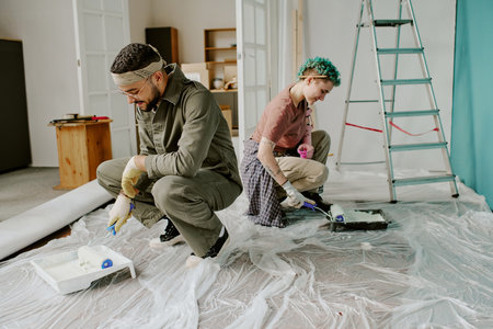 Young adult man and young adult woman kneeling on plastic sheet painting walls with paint rollers during home renovation, ladder and paint trays visible in backgroundの写真素材