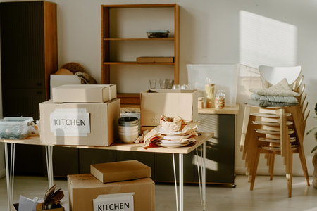 Cardboard boxes labeled kitchen stacked on table with unpacked dishes and kitchenware in bright room, young couple moving and preparing for renovations, no people visibleの写真素材