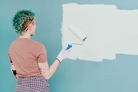 Young adult woman painting wall with paint roller during home renovation, wearing gloves and holding roller while standing in front of partially painted blue wallの写真素材