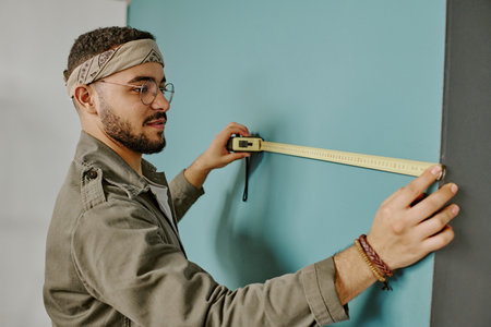 Young adult man measuring wall with tape measure during home renovation, focusing on accuracy while...