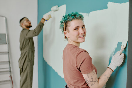 Portrait of young adult Caucasian woman smiling at camera while painting wall with roller, young adult man with beard and glasses painting wall in background during home renovationの写真素材