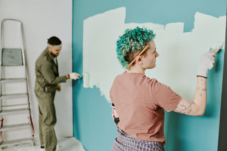 Young adult Caucasian woman with short curly hair painting wall with roller while young adult man...