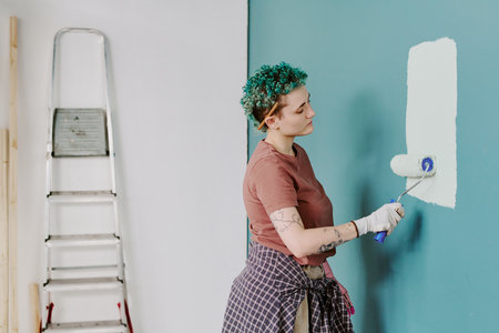 Young adult woman painting wall with roller brush during home renovation, standing near ladder, wearing gloves, focused on applying fresh coat of paintの写真素材