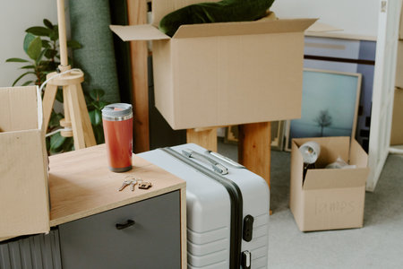 Unpacked cardboard boxes, suitcase, travel mug, and keys on cabinet suggesting young couple moving into new home and preparing for renovations with household items scattered aroundの写真素材