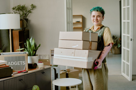 Portrait of young adult woman with short curly hair and prosthetic arm smiling while carrying stack of cardboard boxes during movingの写真素材