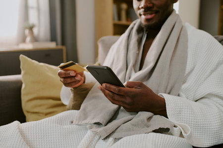 Young adult Black man sitting on sofa holding smartphone and credit card, smiling while making online purchase, engaging in digital shopping from homeの写真素材