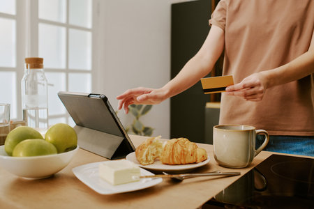 Caucasian young adult woman holding credit card and using tablet for online shopping in kitchen, left hand touching screen while standing near plate with croissants and cup of coffeeの写真素材