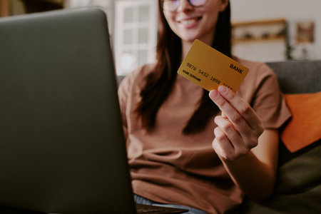 Caucasian young adult woman sitting indoors holding credit card in right hand while using laptop for online shopping smiling slightlyの写真素材
