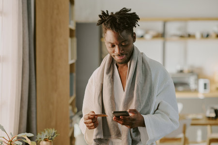 Young adult Black man standing indoors holding credit card and smartphone, smiling while making...