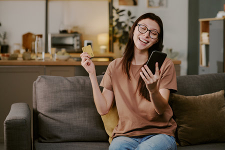 Caucasian young adult woman sitting on sofa holding smartphone and credit card smiling while making online purchase in home settingの写真素材