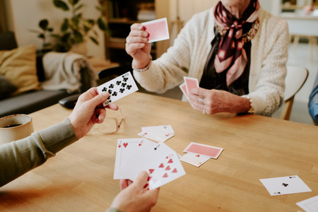 Senior Caucasian woman and middle aged Caucasian man playing card game at wooden table, holding playing cards in hands, interacting and focusing on game, coffee mug on tableの写真素材