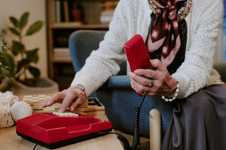 Senior Caucasian woman sitting in living room holding red landline telephone receiver in one hand...