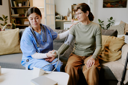Young adult Hispanic woman measuring blood pressure of middle aged woman using digital monitor, sitting together on sofa in home setting, nurse supporting patient armの写真素材