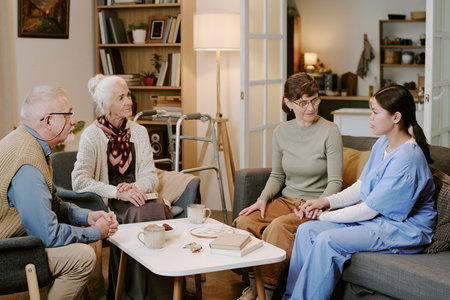 Senior Caucasian man and senior Caucasian woman sitting with middle aged Caucasian woman and young adult Hispanic woman in medical scrubs holding hands, engaging in supportive conversationの写真素材