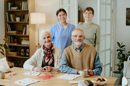 Senior Caucasian woman and senior Caucasian man sitting at table playing board game, young adult Hispanic woman and middle aged Caucasian woman standing behind smiling at cameraの写真素材