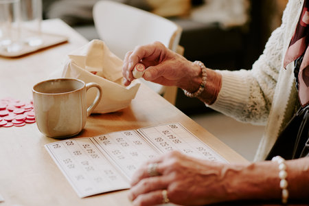 Senior Caucasian woman sitting at table playing bingo, holding numbered bingo chip in hand, marking bingo card, wearing bracelet and sweater, coffee mug on tableの写真素材