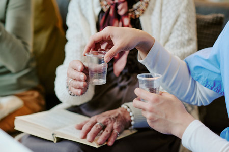 Senior woman sitting with open book receiving plastic cup of water from young adult woman in medical uniform, hands visible, focus on caregiving interactionの写真素材