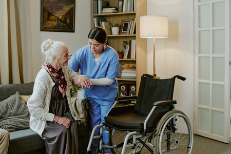 Senior Caucasian woman sitting on sofa being assisted by young adult Hispanic female nurse standing...