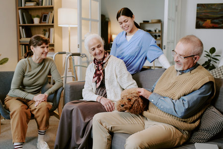 Senior Caucasian woman sitting on sofa smiling at senior Caucasian man petting dog, while young Hispanic woman in nurse uniform standing behind them and middle aged Caucasian woman sitting nearbyの写真素材