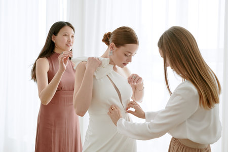 Caucasian young adult woman standing while two young adult women assisting with wedding dress fitting, preparing for wedding celebration in bright room with sheer curtainsの写真素材