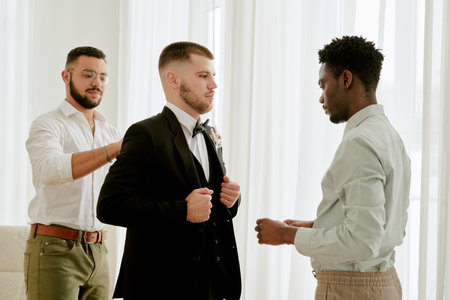 Caucasian young adult man standing in suit being assisted by Black young adult man and young adult man preparing for wedding celebration indoorsの写真素材