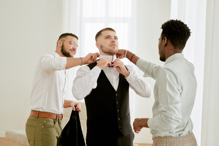 Young adult Caucasian man adjusting bow tie with assistance from young adult Caucasian man and young adult Black man preparing for wedding celebrationの写真素材