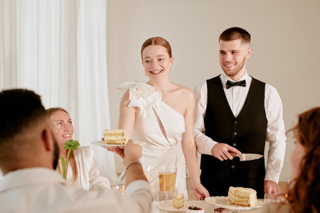 Caucasian young adult bride smiling while serving wedding cake to guest, standing beside Caucasian...